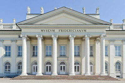 Auf dem Bild ist die Fassade des Museum Fridericianum in Kassel, ein klassizistisches Gebäude mit imposanten Säulen, großen Fenstern und der Aufschrift "Museum Fridericianum", zu sehen. Klarer blauer Himmel im Hintergrund.