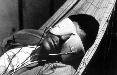 Filmstill das einen Mann mit einer Maske mit Kabeln zeigt.