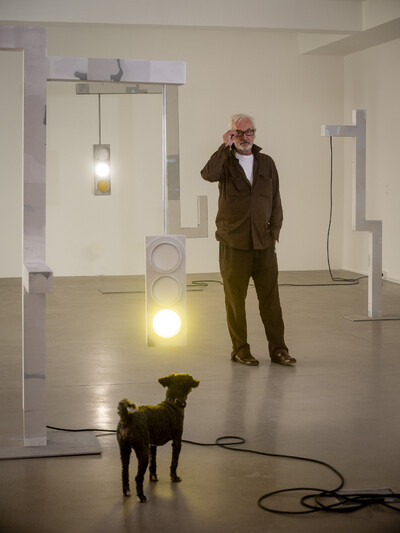 Foto von Peter Fischli, umgeben von seiner Kunst und einem Hund in einem Ausstellungsraum.