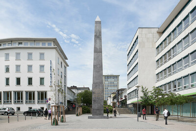Ein hoher, grauer Obelisk steht in der Stadt Kassel. Auf dem Obelisken steht ein Satz über Gastfreundschaft.