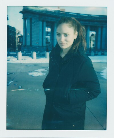 Analog-Foto von Rachel Rose. Sie steht vor einem alten Gebäude. Sie hat einen Mantel an.