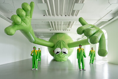 Vier Menschen in Froschkostümen halten gemeinsam einen riesigen Kermit-Luftballon in einem Museum fest.