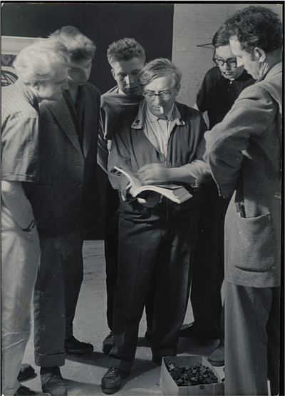 Arnold Bode steht mittig im Raum und hält ein aufgeschlagenes Buch, er wird von fünf Männern umrundet