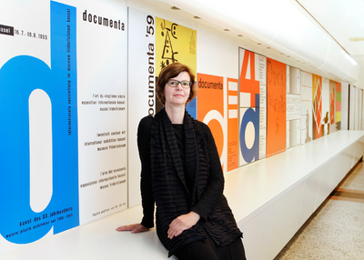 Die Direktorin vor einer Wand mit Ausstellungsplakaten der documenta.