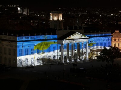 Das Fridericianum bei Nacht, auf dessen Fassade eine farbige Videoprojektion mit blauen, weißen und gelben Elementen zu sehen ist; Menschen sind vereinzelt auf dem dunklen Vorplatz erkennbar.