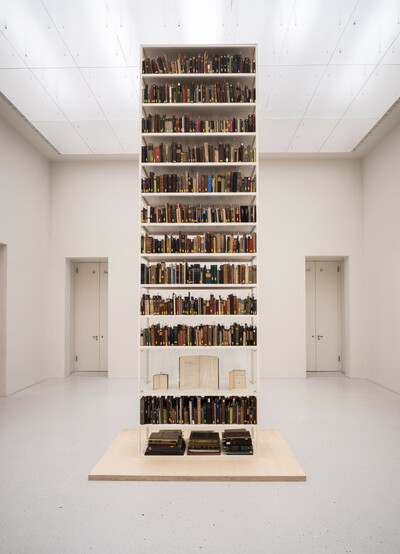 Regalhoher, freistehender Bücherschrank in einem weißen, minimalistischen Raum. Die Regalböden sind mit zahlreichen alten Büchern gefüllt, einige Bücher und Dokumente sind in einer Vitrine im unteren Bereich gesondert ausgestellt.