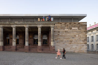 Das damalige Modekaufhaus Sinn-Leffers ist abgebildet, die Figurengruppe "Die Fremden" von Thomas Sch&uuml;tte ist auf dem Dach des Portikus zu sehen, im Vordergrund laufen zwei Menschen am Geb&auml;ude vorbei