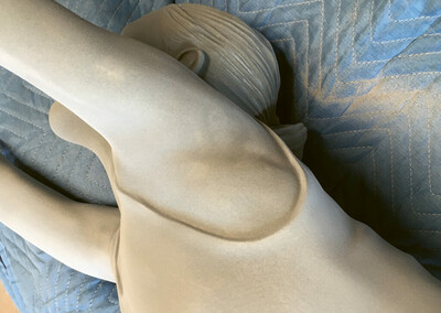 Nahaufnahme einer beigen Skulptur mit realistisch geformtem Oberkörper und erhobenem Arm, liegend auf einer wattierten blauen Decke mit Zickzacknähten.