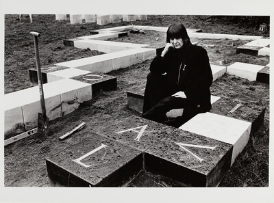 Eine Frau in schwarzer Kleidung sitzt auf den steinernen Quadern eines minimalistischen Monuments