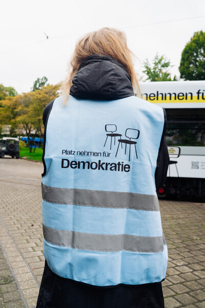 Person from behind, wearing a vest. On the webste it says "Platz nehmen für Demokratie".
