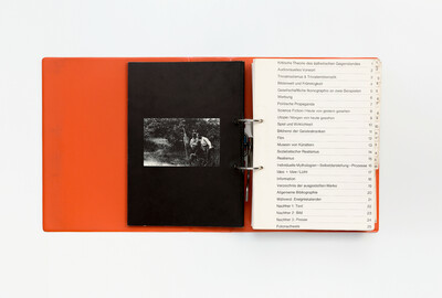 Doppelseite aus dem Ausstellungskatalog der documenta 5 von 1972 in Ringbuchform. Links ein schwarzes Blatt mit eingeklebtem Schwarz-Weiß-Foto, das zwei Männer in ländlicher Umgebung zeigt. Rechts das tabellarisch aufgebaute Inhaltsverzeichnis mit 25 nummerierten Kapiteln, darunter Themen wie „Politische Propaganda“, „Science Fiction / Heute von gestern gesehen“, „Museen von Künstlern“ oder „Individuelle Mythologien – Selbstdarstellung – Prozesse“.