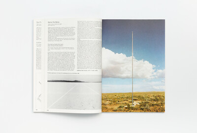 Doppelseite aus dem Ausstellungskatalog der documenta 6 mit einem Text zu Walter De Maria und einem Farbfoto seines Werks „The Vertical Earth Kilometer“. Das Bild zeigt eine karge Landschaft mit blauem Himmel und einer senkrecht aus dem Boden ragenden Metallstange als sichtbares Zeichen der im Erdreich versenkten Skulptur.