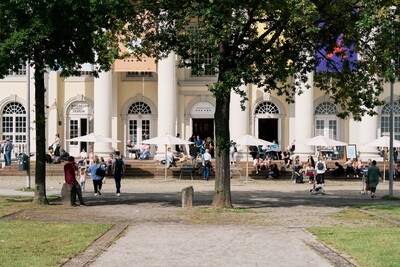 Außenansicht des Cafés im Fridericianum, mit Bäumen im Vordergrund und dem Außenbereich des Cafés im Hintergrund. Es sind viele Menschen zu sehen.