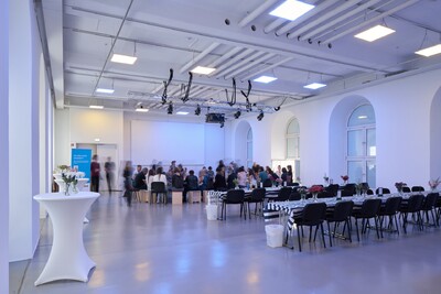 Veranstaltungssaal im Fridericianum mit einer Reihe von Sthlen und Tischen, sich bewegenden Menschen und einer weißen Leinwand am Ende des Raumes.