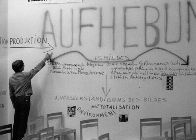 Wand mit Text. Links steht eine Person und sprüht mit einer Farbdose einen Pfeil an die Wand.