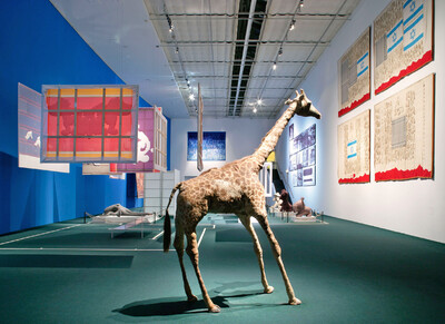 Exhibition view with stuffed giraffe and colorful geometric objects in a blue room.