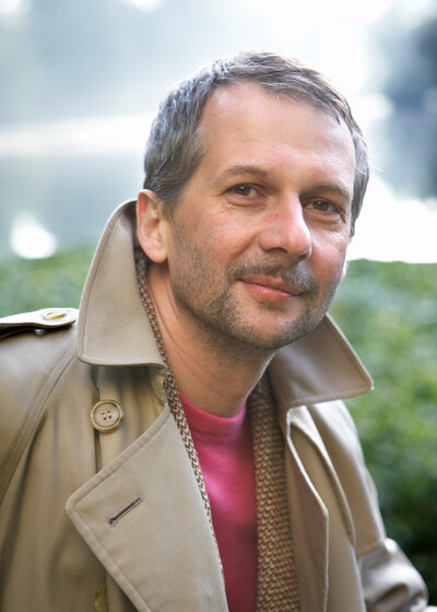Portrait of the Artistic Director with open trench coat and pink sweater outdoors.