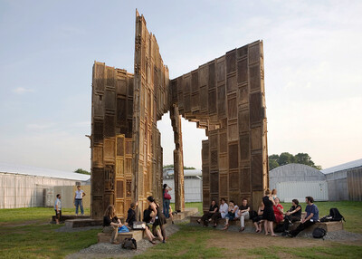Large wooden construction made of old doors and windows, set up outdoors, people sitting underneath.