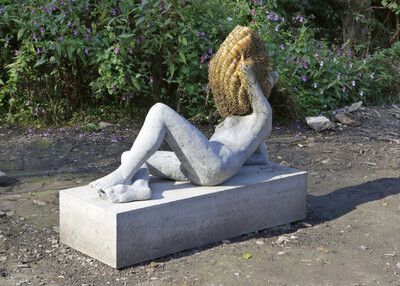Eine steinerne Skulptur einer liegenden, nackten menschlichen Figur liegt auf einem Betonsockel. Ihr Kopf ist durch einen großen Bienenkorb ersetzt. Im Hintergrund wachsen grüne Pflanzen mit violetten Blüten.