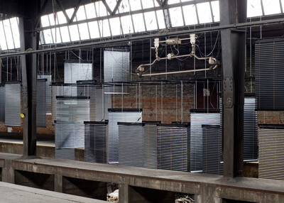 Mehrere Metalljalousien hängen in unterschiedlichen Höhen in einer verlassenen Industriehalle. Die Konstruktion wirkt wie eine Installation, während altes Mauerwerk und rostige Stahlträger die Umgebung bestimmen.