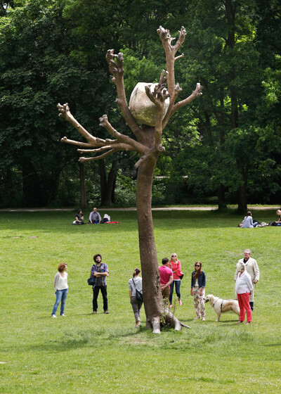 Eine große Skulptur eines kahlen Baumes mit einem massiven Stein in seiner Astgabel steht in einer grünen Parklandschaft. Mehrere Menschen und ein Hund befinden sich in der Nähe.