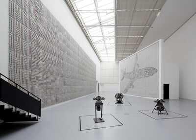 Eine Wand in der documenta Halle zeigt ein strukturiertes Muster, während eine andere ein großes Flugzeugmotiv trägt. Drei Maschineninstallationen stehen auf dem Boden.