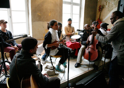 Eine Musikgruppe spielt in einem Raum mit abgeblätterten Wänden. Eine Person mit einer kleinen Gitarre sitzt im Zentrum, umgeben von anderen Musikern mit verschiedenen Instrumenten, darunter ein Cello, eine Gitarre und Schlagzeug.