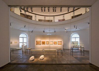 Innenansicht einer Museumsausstellung mit Glaswand im Vordergrund. An der beleuchteten Rückwand sind gerahmte Kunstwerke aufgehängt, während Ausstellungstische und Skulpturen im Raum verteilt sind.