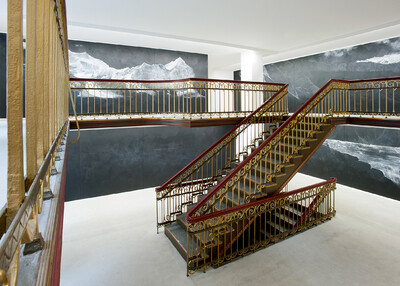 Eine imposante Treppe mit goldenen Geländern windet sich in einem weißen Raum. An den Wänden sind großformatige schwarz-weiße Landschaftsgemälde zu sehen.