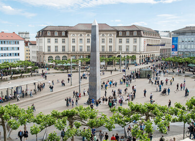 Das Foto zeigt den Königsplatz in Kassel mit dem Obelisken von Olu Oguibe im Zentrum, umgeben von zahlreichen Besuchenden.