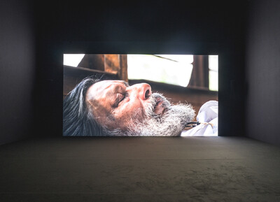 Nahaufnahme eines Mannes mit grauem Bart der die Augen geschlossen hat, präsentiert auf einem großformatigen Screen in einem dunklen Ausstellungsraum.