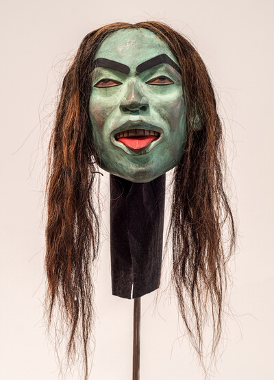 Detailaufnahme einer geschnitzten rituellen Maske mit grüner Bemalung und langen Haaren.
