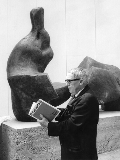 Arnold Bode steht vor einer Statue und liest im Katalog der documenta 3.