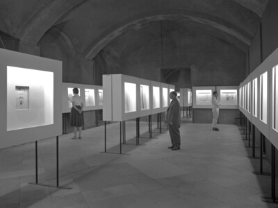 Ausstellung in einem Gewölbe. Man sieht drei Personen die sich ausgestellte Kunst ansehen. Diese ist in weißen Schaukästen präsentiert.