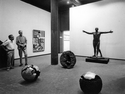 Zwei Männer stehen in einem Ausstellungsraum. Zu sehen sind mehrere Skulpturen auf dem Boden und im Raum verteilt stehend sowie ein Kunstwerk an der Wand hängend.