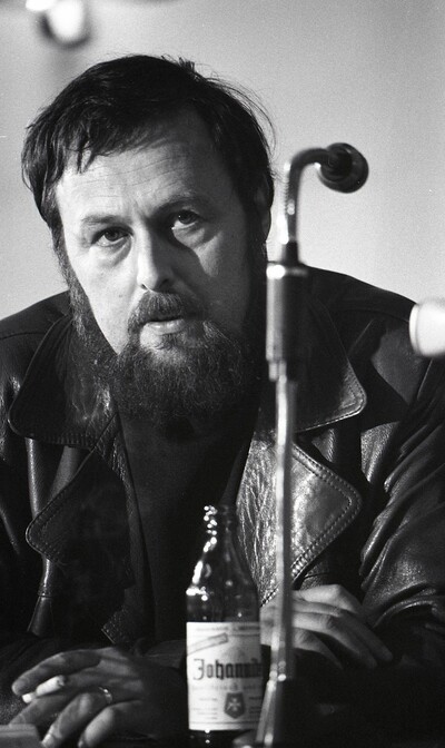 Schwarzweiß-Fotografie von Harald Szeemann. Er trägt einen Bart. Vor ihm steht ein Bier, in der Hand hält er eine brennende Zigarette.