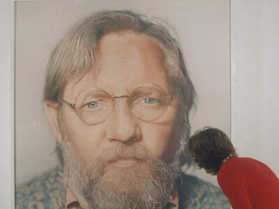 Eine Person sieht sich ein großes Bild an. Das Bild zeigt einen Männerkopf mit Brille und Bart.