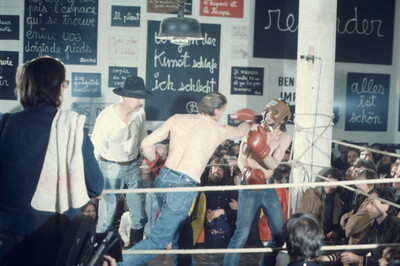 Ein Boxring mit mehreren Personen darin und drumherum. Zwei davon sind Oberkörperfrei, tragen Boxhandschuhe und kämpfen.