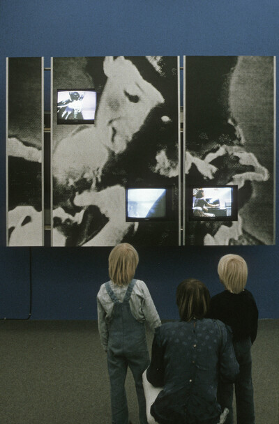 Drei Kinder betrachten eine Videoarbeit mit mehreren Monitoren, eingebettet in ein großformatiges Schwarz-Weiß-Bild an einer blauen Wand.
