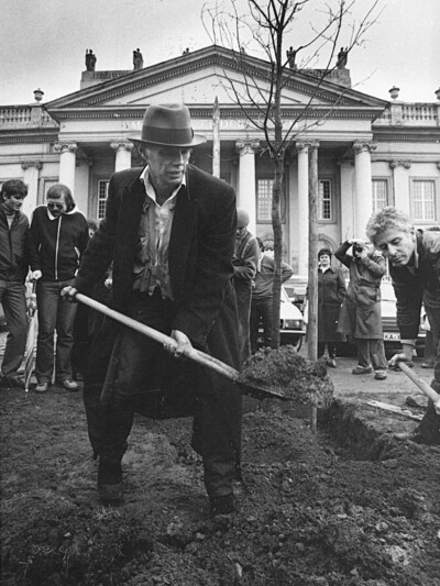 Schwarzweißaufnahme einer Pflanzaktion vor dem Museum Fridericianum in Kassel. Im Vordergrund schaufelt Joseph Beuys Erde, im Hintergrund beobachten Passant*innen die Szene. Die Aktion ist Teil der Arbeit „7000 Eichen“ von Joseph Beuys.