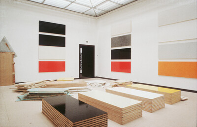 Blick in einen hellen Ausstellungsraum mit geometrisch-abstrakten Farbflächen an den Wänden und gestapelten farbigen Holzplatten am Boden.