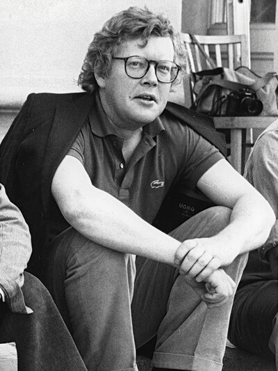 Schwarzweißporträt eines sitzenden Mannes mit Brille, lockigem Haar und Poloshirt. Er hat eine Jacke über die Schultern gelegt und blickt in die Kamera. Im Hintergrund sind Stühle und eine Fototasche zu sehen.
