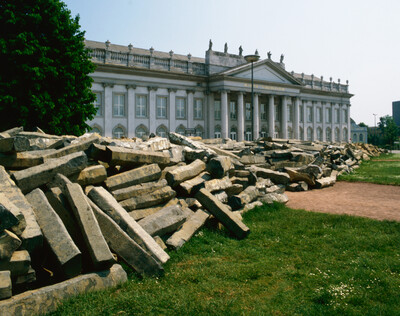 Vor dem Museum Fridericianum in Kassel liegt ein großer Haufen grob behauener Basaltsteine auf einer Wiese. Die Steine sind Teil der Arbeit „7000 Eichen“ von Joseph Beuys.