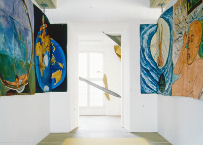 Blick in einen Ausstellungsraum mit großformatigen, farbintensiven Gemälden an den Seitenwänden, die surrealistische Szenen und Weltkarten zeigen. Im Hintergrund ragt ein längliches Objekt diagonal durch einen Türdurchgang.