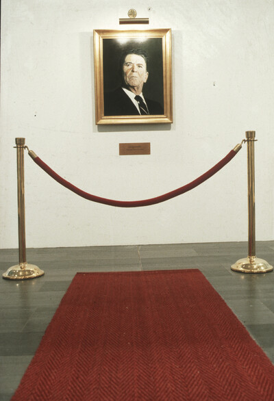 Ein gerahmtes Porträt von Ronald Reagan hängt an einer weißen Wand, davor ein roter Teppich und eine Absperrung mit goldenen Pfosten und rotem Seil.