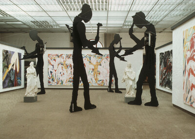 Ausstellungsansicht mit überlebensgroßen, schwarzen Silhouettenfiguren mit mechanischen Elementen, die Werkzeuge halten. Zwischen ihnen stehen klassizistische weiße Skulpturen. An den Wänden hängen großformatige, farbintensive Gemälde.