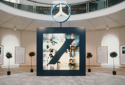 Installation in einer Rotunde mit einem großen, freistehenden blauen Kubus, in dessen Mitte ein unscharfes Foto von demonstrierenden Personen mit einem übergroßen Logo der Deutschen Bank überblendet ist. Darüber schwebt das Mercedes-Stern-Logo. Umgeben ist die Szene von kleinen Topfbäumen und gerahmten Texttafeln an den Wänden.