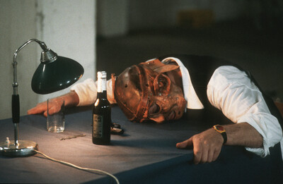 Eine Person mit deformierter Maske liegt mit dem Kopf auf einem Tisch, umgeben von einer Flasche, einem Glas, einer Schere und einer Tischlampe. Die Kleidung wirkt formell, das Licht ist gedämpft. Die Szene erinnert an eine theatralische oder performative Situation.