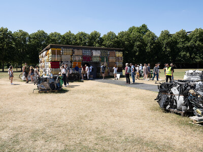 Ein Gebäude aus gepressten Stoffballen steht in der Karlsaue in Kassel. Besuchende betreten das Innere, um das Material und die Konstruktion zu erkunden.