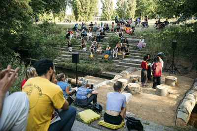 Menschen sitzen auf Stufen am Flußufer und schauen eine Performance an.